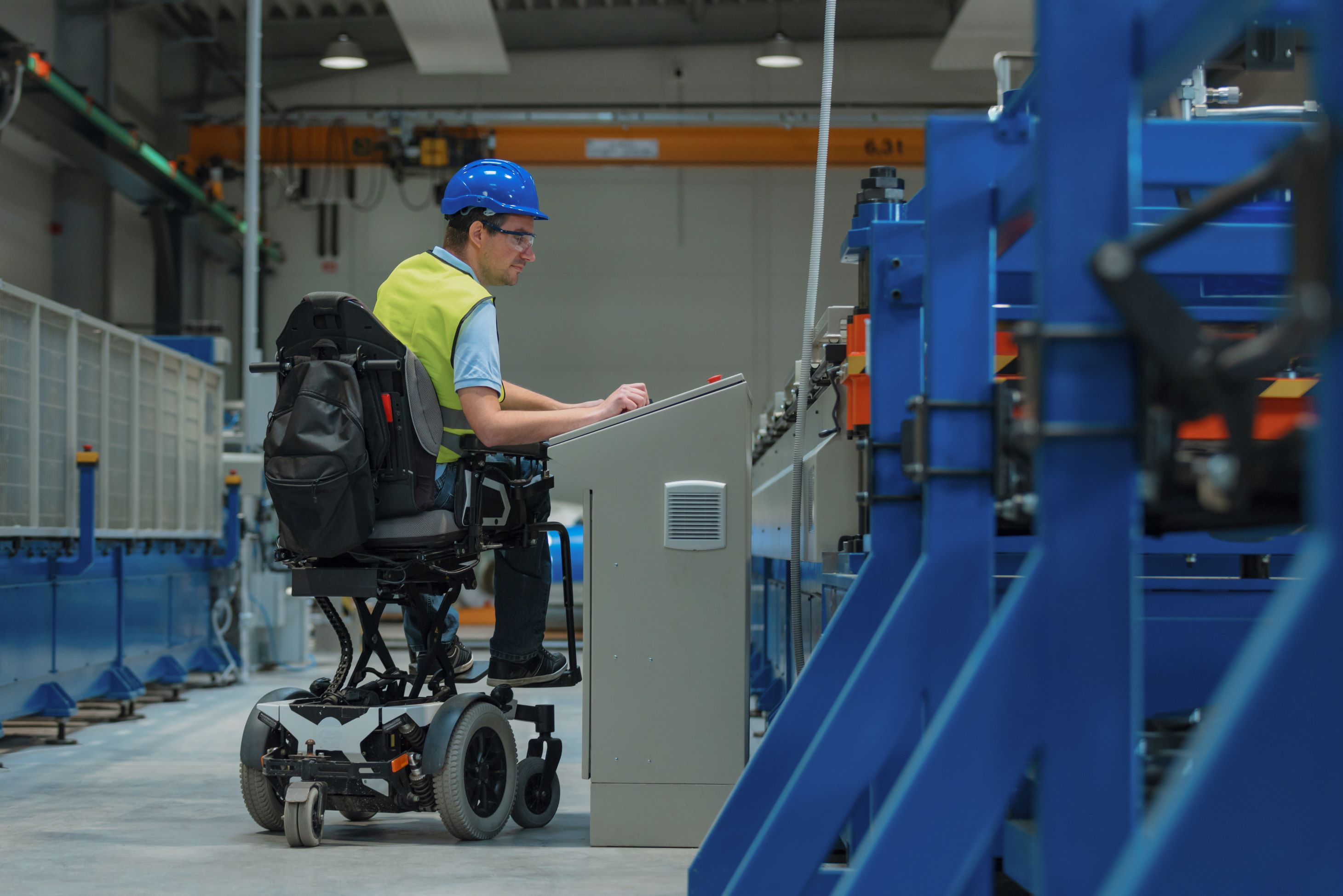 Engineeer who uses a motorized wheelchair at work in factory with yellow vet and blue helmet.